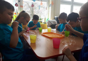 Sześcioro dzieci przy stoliku próbuje osadzić bańki mydlane na stole.